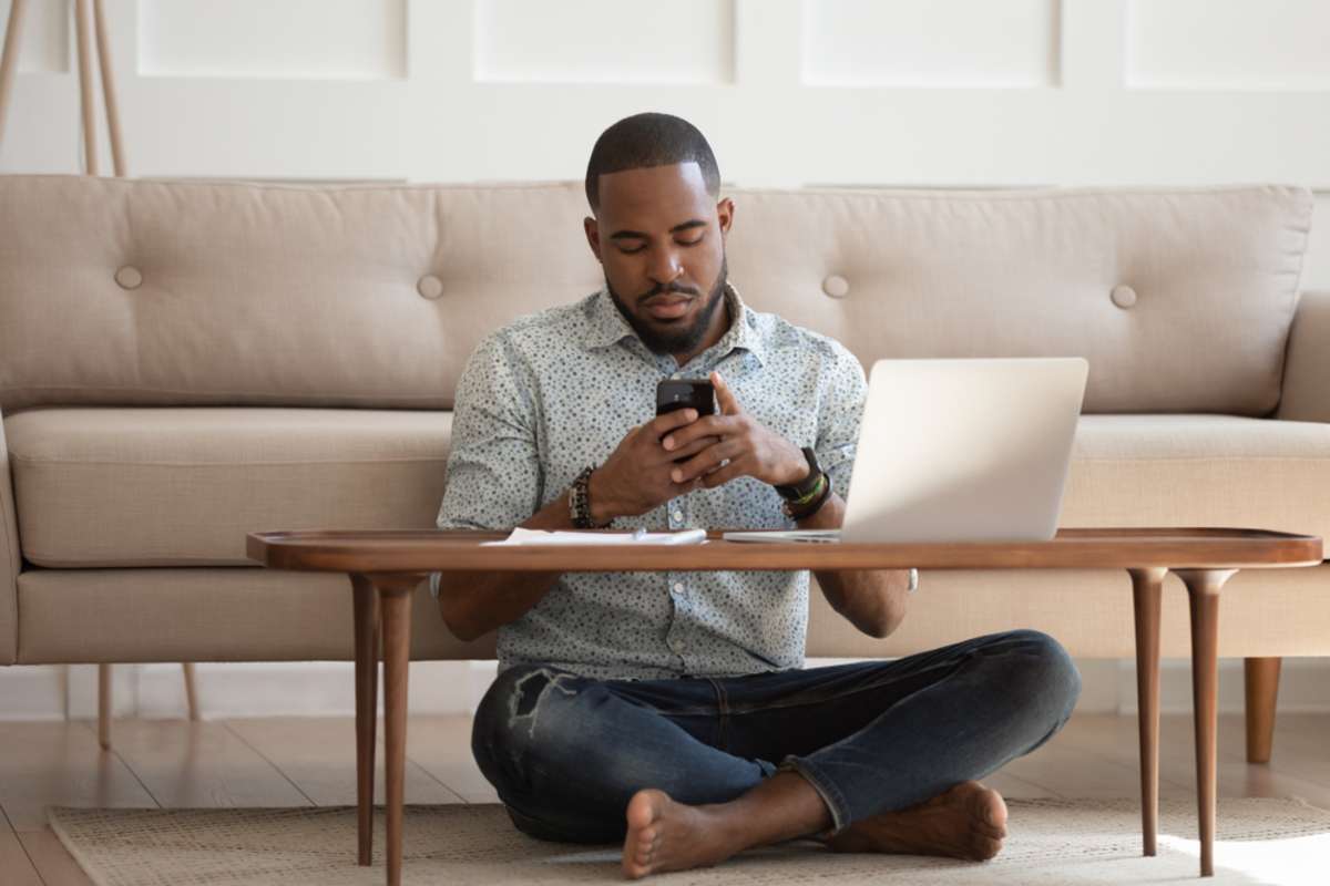 African guy sits leaned on couch cross-legged on carpet near coffee table hold cellphone distracted from work chat online