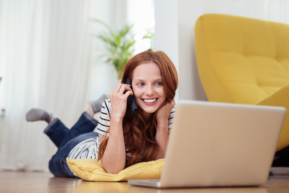 Attractive Young Woman Talking to Someone on Mobile Phone While Relaxing at the Living Room with Laptop Computer