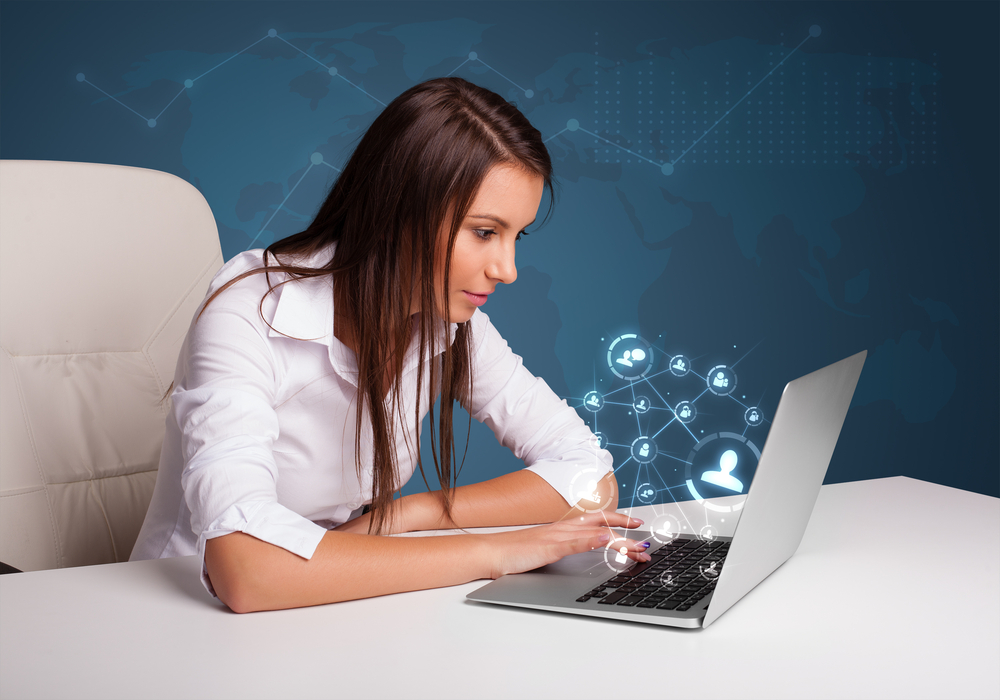 Beautiful young lady sitting at desk and typing on laptop with social network icons coming out.