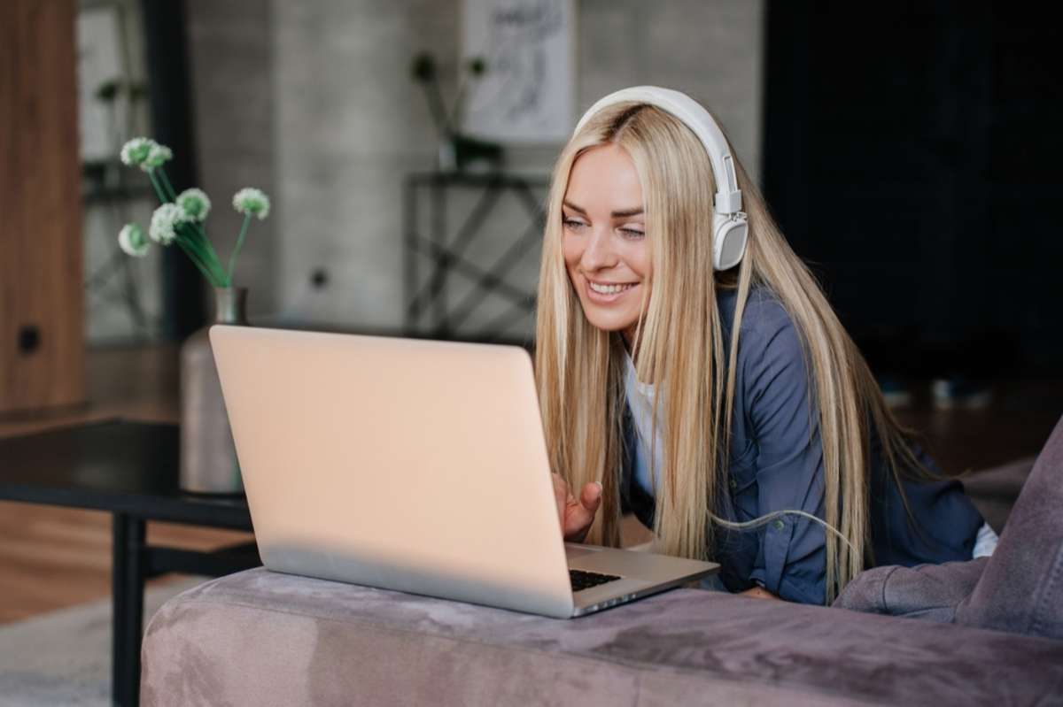 Cheerful blonde young woman in headphones smiles sits on couch with laptop makes video call toothy smiles in adorable mood