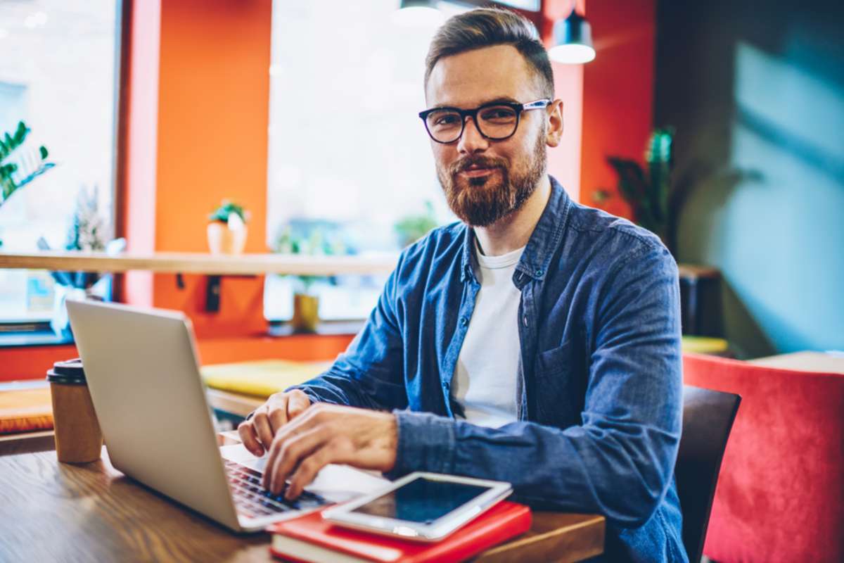 Half length portrait of successful bearded designer smiling at camera while working on freelance at netbook