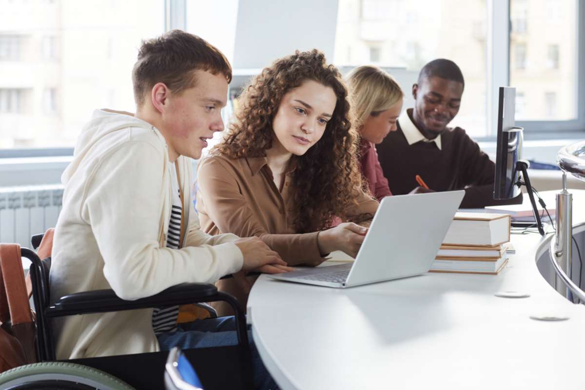 Side view at multi-ethnic group of students using laptop while studying in college, featuring boy using wheelchair