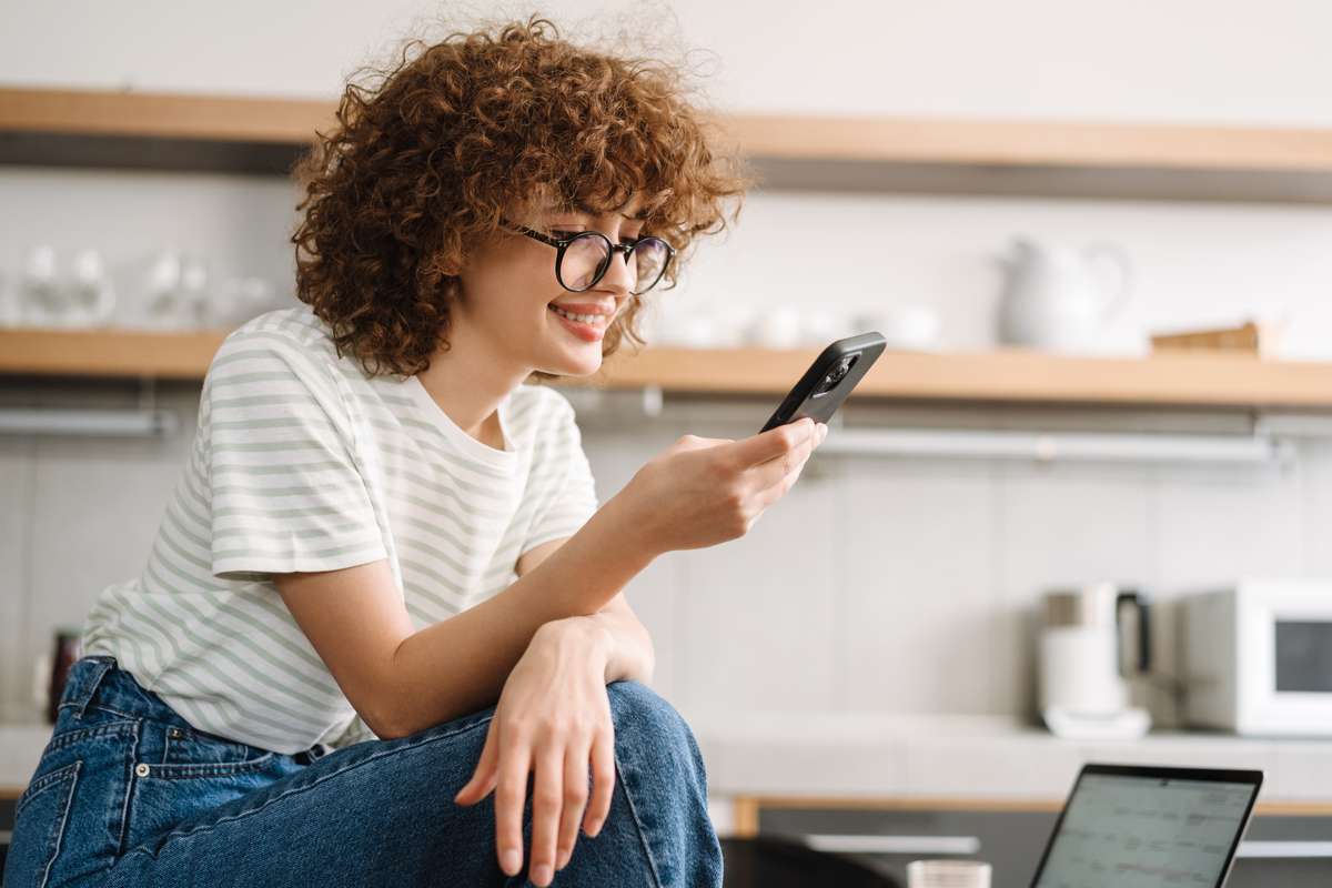 Smiling curly woman using cellphone while sitting on table at kitchen