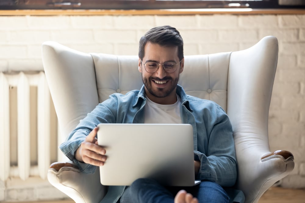 Smiling young man wearing glasses using laptop, sitting in cozy armchair