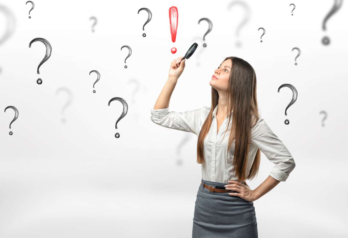 Young businesswoman with magnifying glass over her head looking at red exclamation mark surrounded by question marks