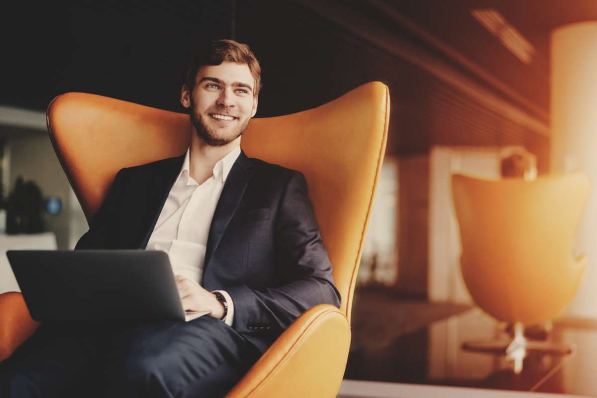 Young smiling successful man entrepreneur in formal business suite with a beard sitting on orange armchair with laptop